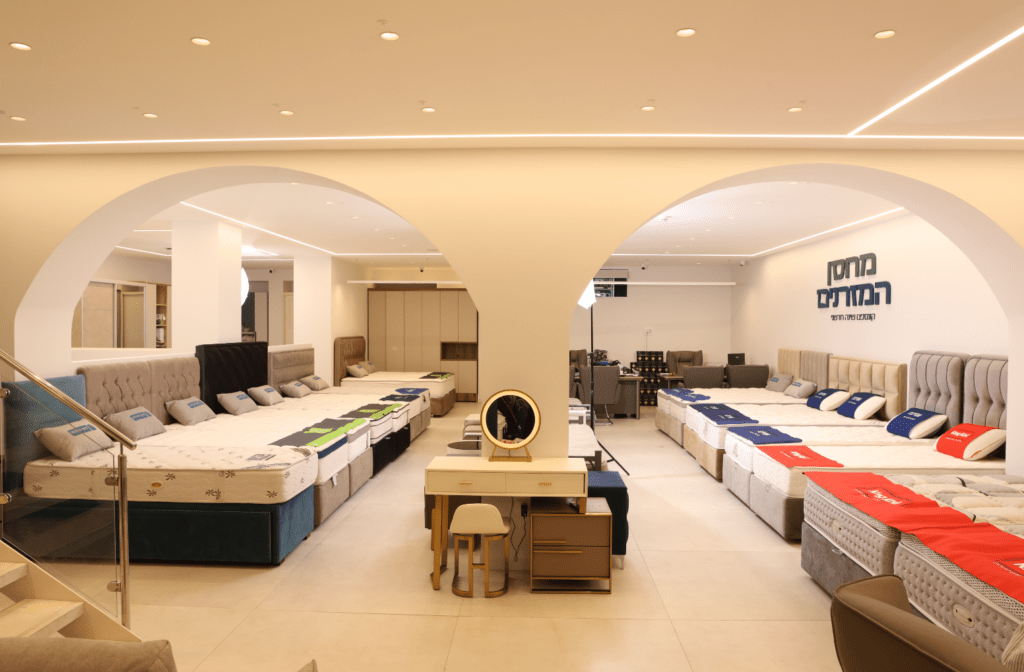 בתוך חנות מרווחת ומוארת, שורות של מיטות ומזרנים מוצגות משני צידי חלל פתוח עם קשתות.