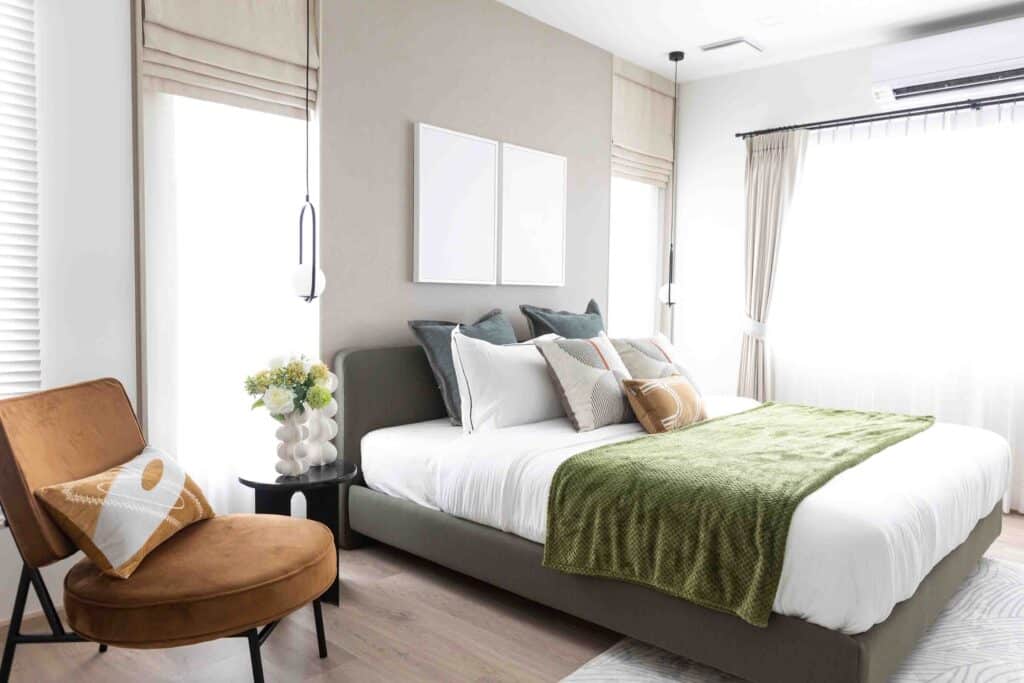 חדר שינה מואר עם מיטה, כריות, שמיכה ירוקה, שידה עם פרחים, וכיסא חום.